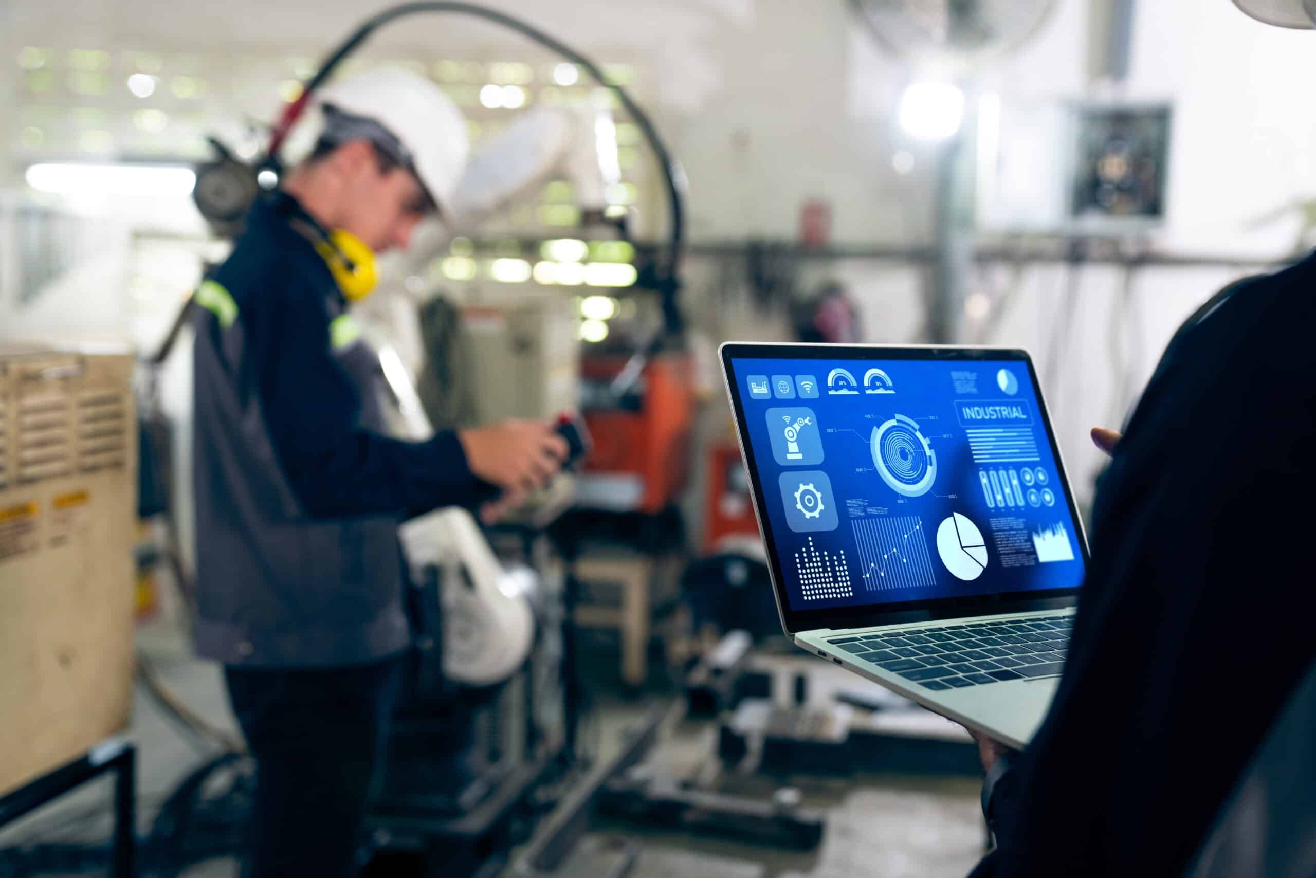 Two factory workers adjusting machinery, one with a laptop showing a software dashboard